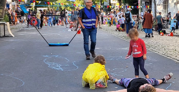 Szenerie eines Straßenfests. Zwei Kinder spielen auf einer Straße mit Kreide, nebendran steht eine Person mit blauer autofrei-Weste